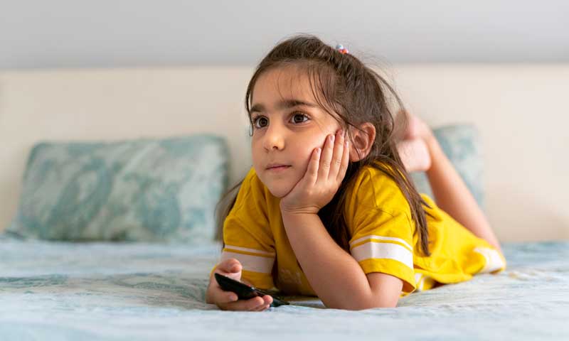 A young girl laying on a bed watching TV with the remote in her hand