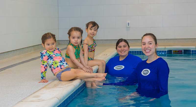 Three young swimming students sitting on the edge of a pool with two Njswim School teachers.